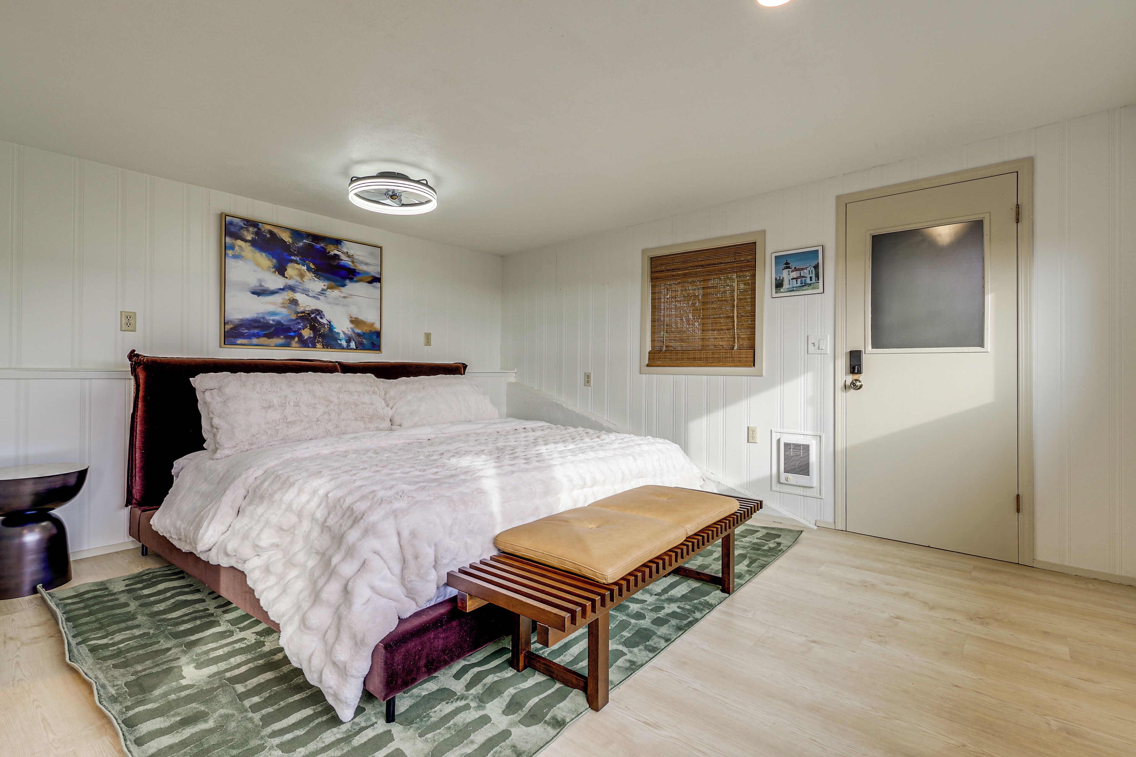 A bedroom with a bed, a bench, a green rug, a wooden bench, a painting, and a door.