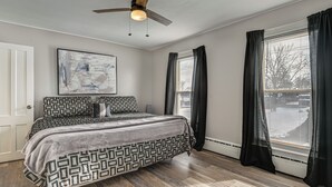 Ein Schlafzimmer mit einem Bett, einem Deckenventilator, einem Fenster mit Vorhängen und einem an der Wand hängenden abstrakten Gemälde.