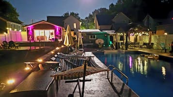 A swimming pool area with lounge chairs and umbrellas, surrounded by buildings with illuminated exteriors.