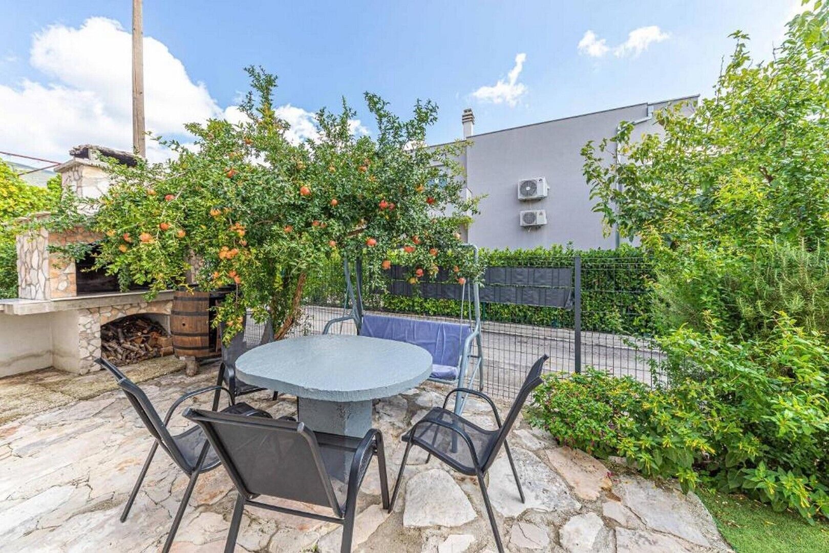 An outdoor patio with a stone table and chairs, surrounded by greenery and a brick wall.