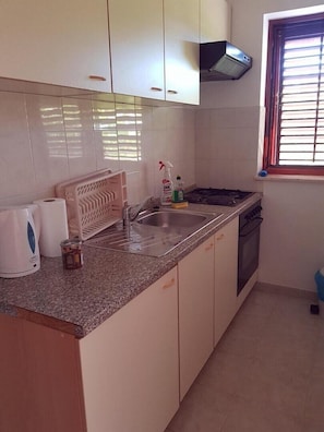 A kitchen with white cabinets, a stainless steel sink, a gas stove, and a window with blinds. - Two Bedroom Apartment (Kaštel Štafilić)
