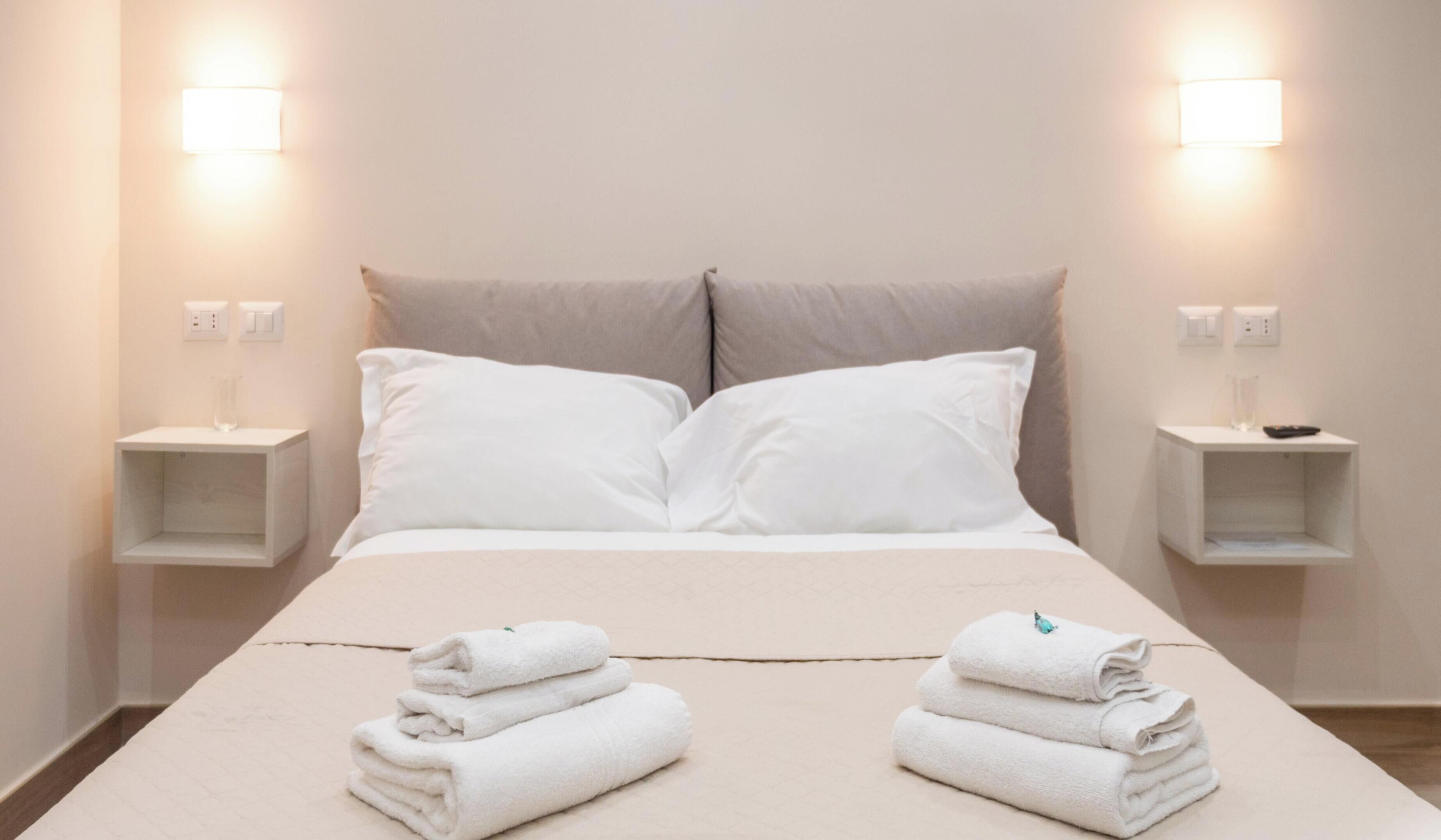 A neatly made bed with two pillows, two bedside tables with lamps, and two rolled towels on the bed.