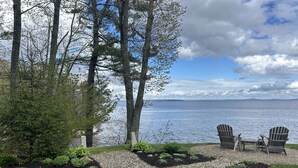 Ein Pfad führt zu zwei Adirondack-Stühlen am See, umgeben von Bäumen und bewölktem Himmel.