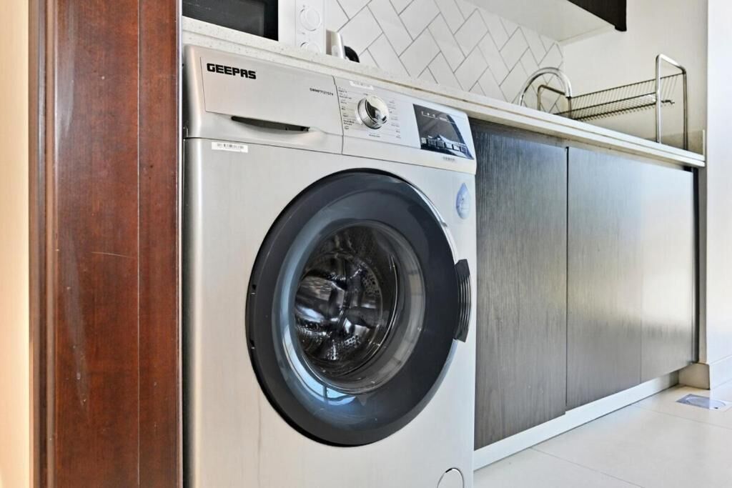 Vue de face d’un lave-linge à chargement frontal de la marque GEEPAS, installé dans une cuisine moderne avec un dosseret de carreaux blancs et des armoires en acier inoxydable.