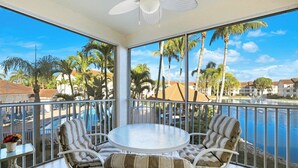 A balcony with a round glass table and striped wicker chairs, overlooking a swimming pool and palm trees. - St. Croix at Pelican Marsh (Naples)