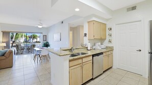 A modern kitchen with granite countertops, wooden cabinets, and a dishwasher. There is a dining area with a table and chairs, and a balcony with a view of palm trees. - St. Croix at Pelican Marsh (Naples)