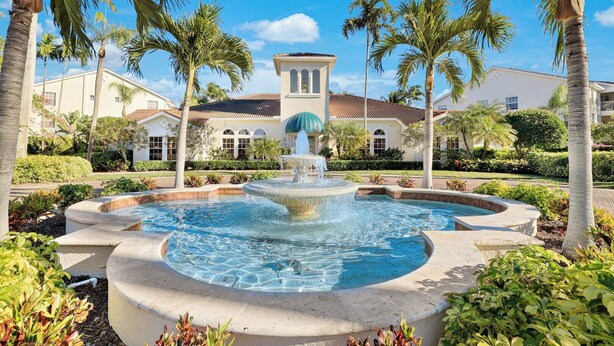 A circular fountain with water jets in the center, surrounded by palm trees and a building with a tiled roof in the background. - St. Croix at Pelican Marsh (Naples)