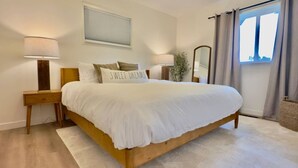 A neatly made bed with a white comforter and pillows, a bedside table with a lamp, a mirror, and a window with curtains. - 3BR Lacey home with bonus room, workspace, fenced yard & patio near trail, shops, and I-5. (Lacey)