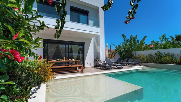 Uma casa moderna com piscina, um alpendre com cadeiras de descanso e vegetação exuberante.