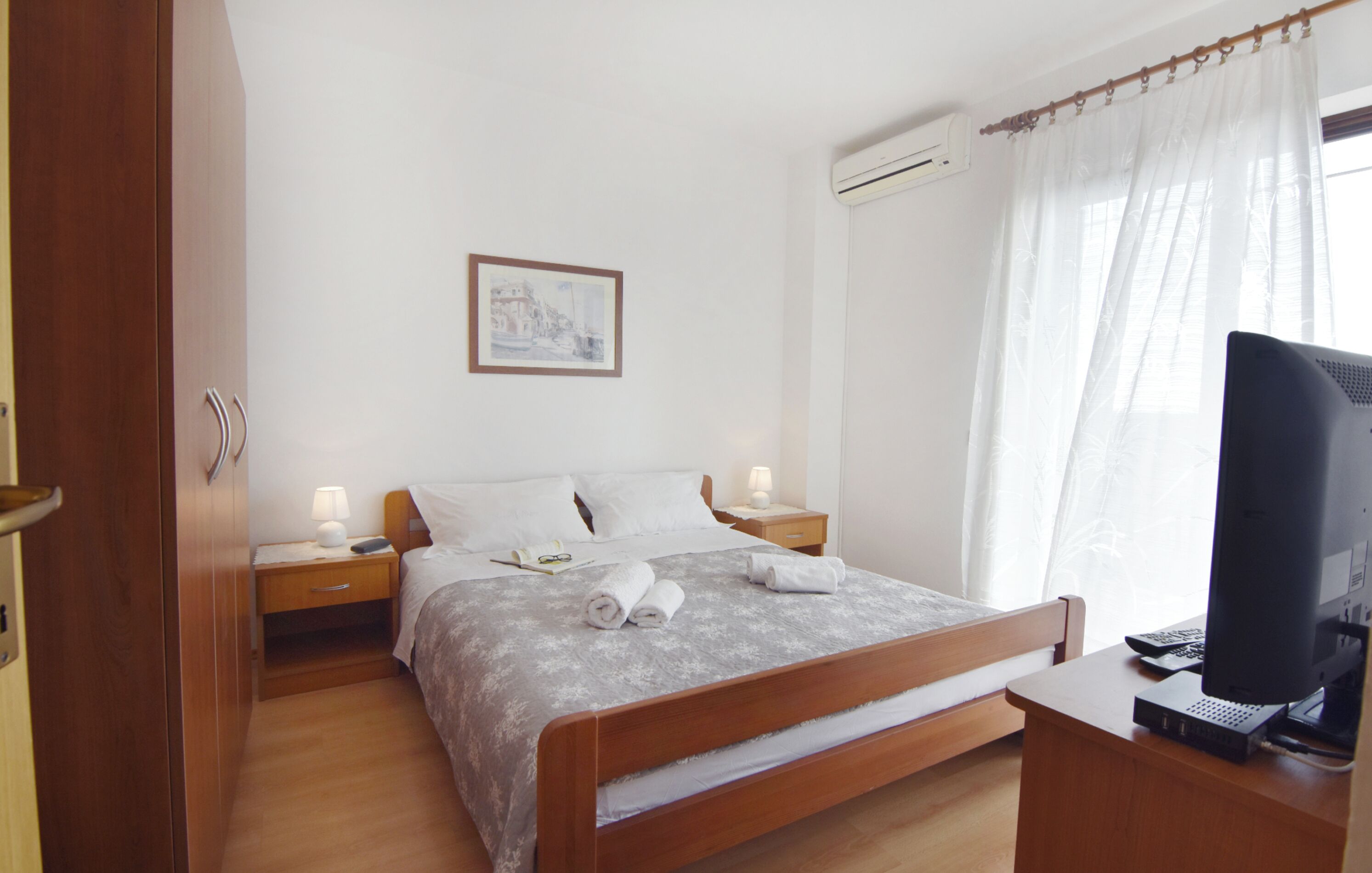 Een net opgemaakt bed met kussens en een deken, een nachtkastje met een lamp, een ingelijst schilderij aan de muur en een bureau met een beeldscherm.