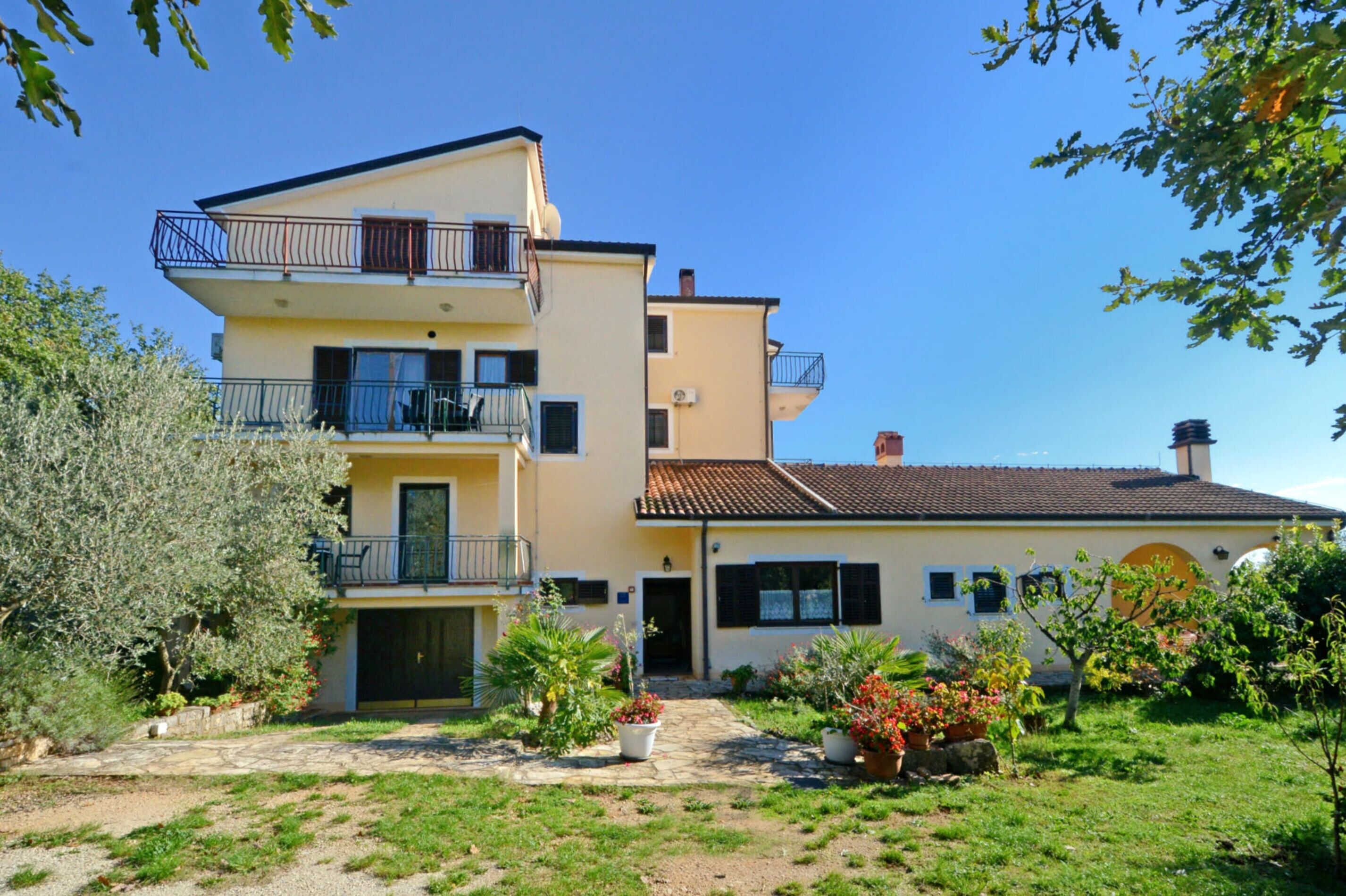 A multi-story residential building with balconies, a garden with potted plants, and a clear blue sky.
