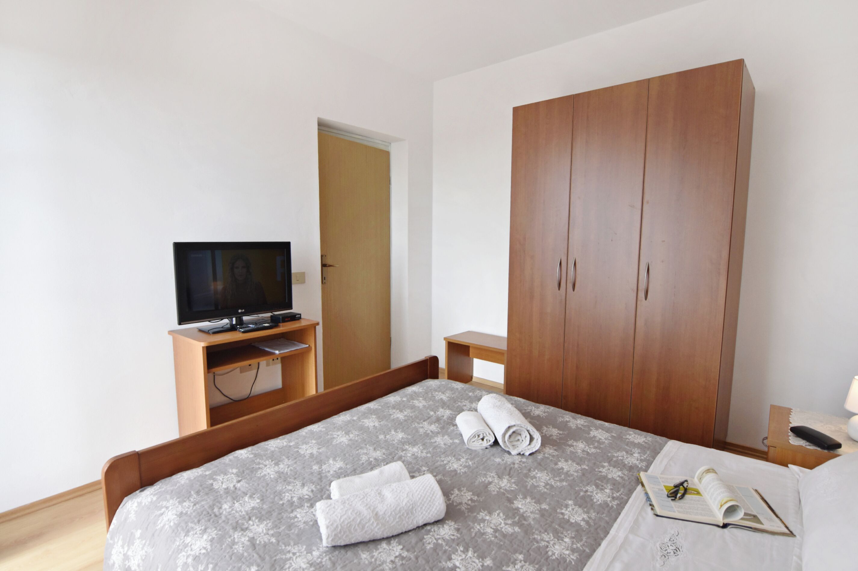 A hotel room with a bed, a television on a wooden stand, a wardrobe, and a small bedside table with a lamp.