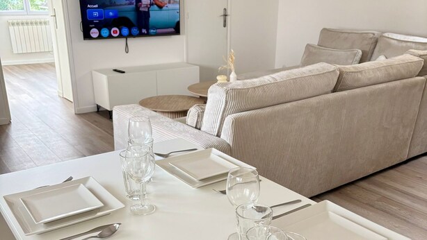 Une table à manger élégamment dressée, avec de l’argenterie, des couverts et des verres, un canapé beige en arrière-plan et un téléviseur diffusant un paysage.