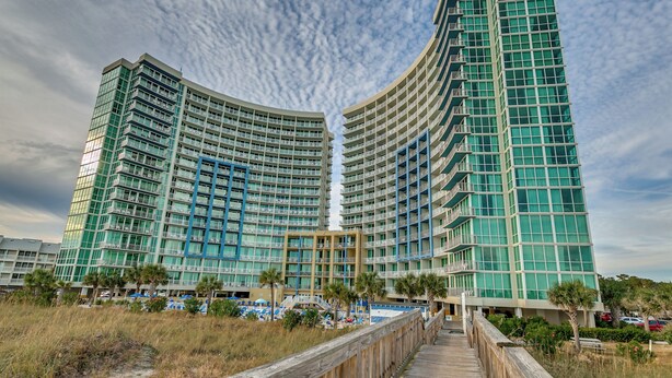 Un hotel moderno de altura con fachada curva, varios balcones y un paseo marítimo que conduce a una zona de piscina.