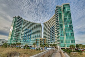 A high-rise hotel with a curved facade, multiple balconies, and a boardwalk leading to a pool area. - Oceanfront, great resort amenities + Free Attraction Tickets! (North Myrtle Beach)