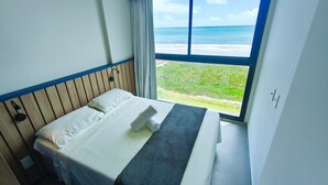 A hotel room with a bed, a window with a view of the ocean, and a wooden headboard. - 02 Quartos no Nomar Carneiros Ecoliving (Tamandaré)