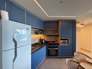 A modern kitchen with blue cabinets, a white refrigerator, a built-in oven, and a dining area with a table and chairs. - 02 Quartos no Nomar Carneiros Ecoliving (Tamandaré)