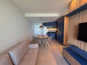 Ein modernes Wohnzimmer mit einer Couch, einem Esstisch und Stühlen. Im Hintergrund ist eine Küche mit blauen Schränken und einem an der Wand befestigten Fernseher zu sehen.