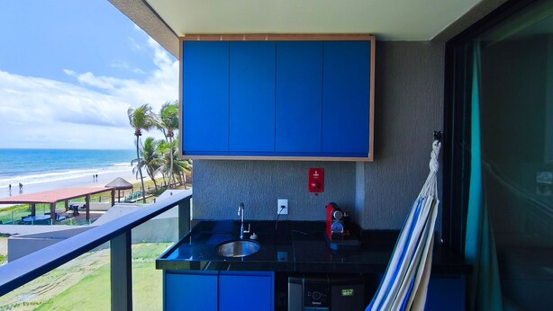 A balcony with a view of the beach, a sink, and a red fire alarm. - 02 Quartos no Nomar Carneiros Ecoliving (Tamandaré)