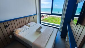 A bedroom with a bed, a window with a view of the beach, and a blue wall. - 02 Quartos no Nomar Carneiros Ecoliving (Tamandaré)