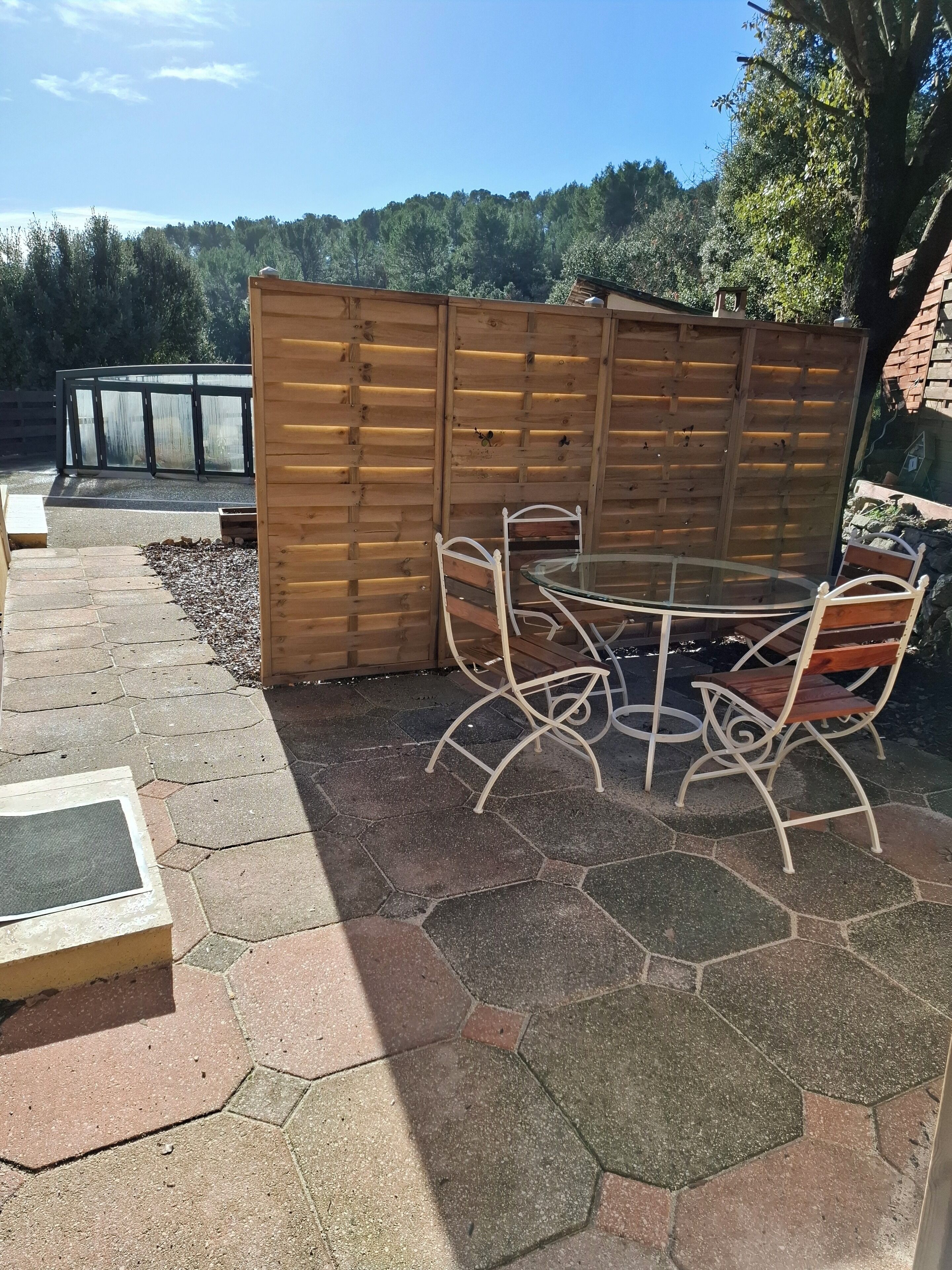 Un patio al aire libre con suelo de piedra hexagonal, valla de madera y una mesa redonda de vidrio con sillas de metal.