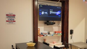 A room with a wooden table and chairs, a television mounted on the wall, and a shelf with various items. - Treehouse Hostel (Montreal)