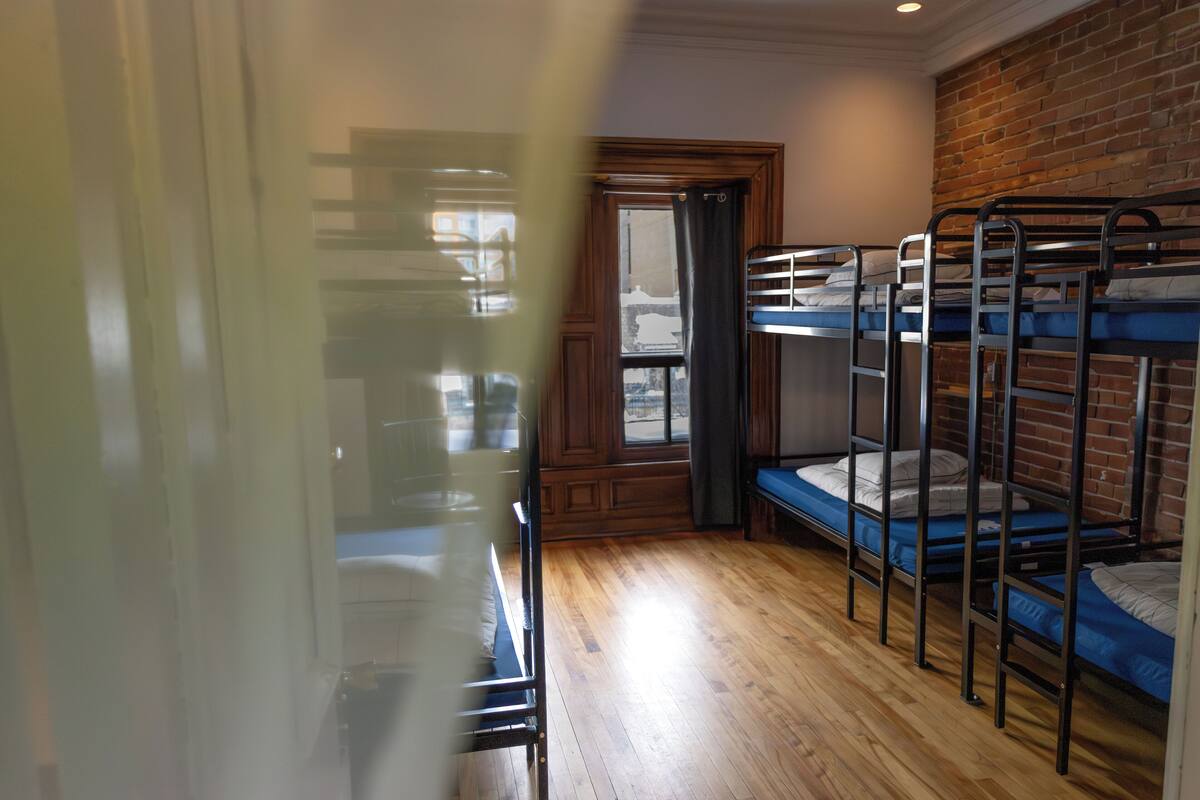 A room with bunk beds, wooden flooring, a brick wall, and a window with curtains.