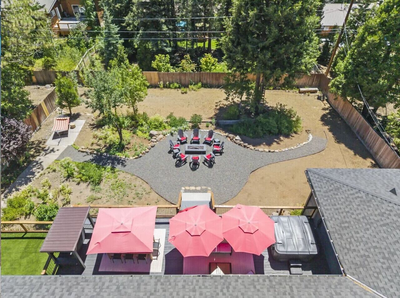 Une vue aérienne d’une cour arrière avec un foyer entouré de chaises et de tables, une terrasse en bois avec des parasols et une zone délimitée par une clôture, où se trouvent des arbres.