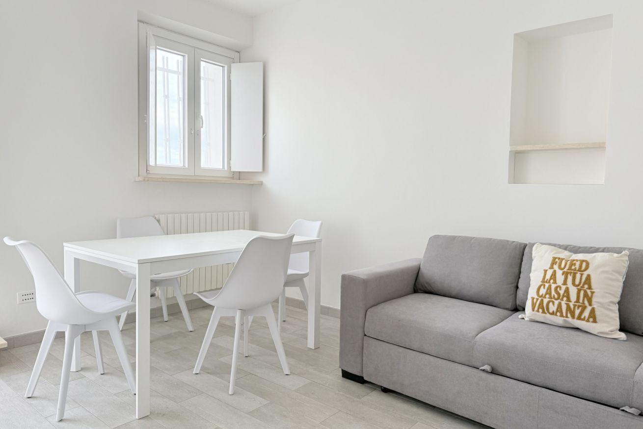 A room with a white dining table, four chairs, a grey sofa, and a window with open blinds.