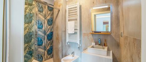 A bathroom with a bidet, a towel rack, a shower curtain with a leaf pattern, and a mirror with a light above it.