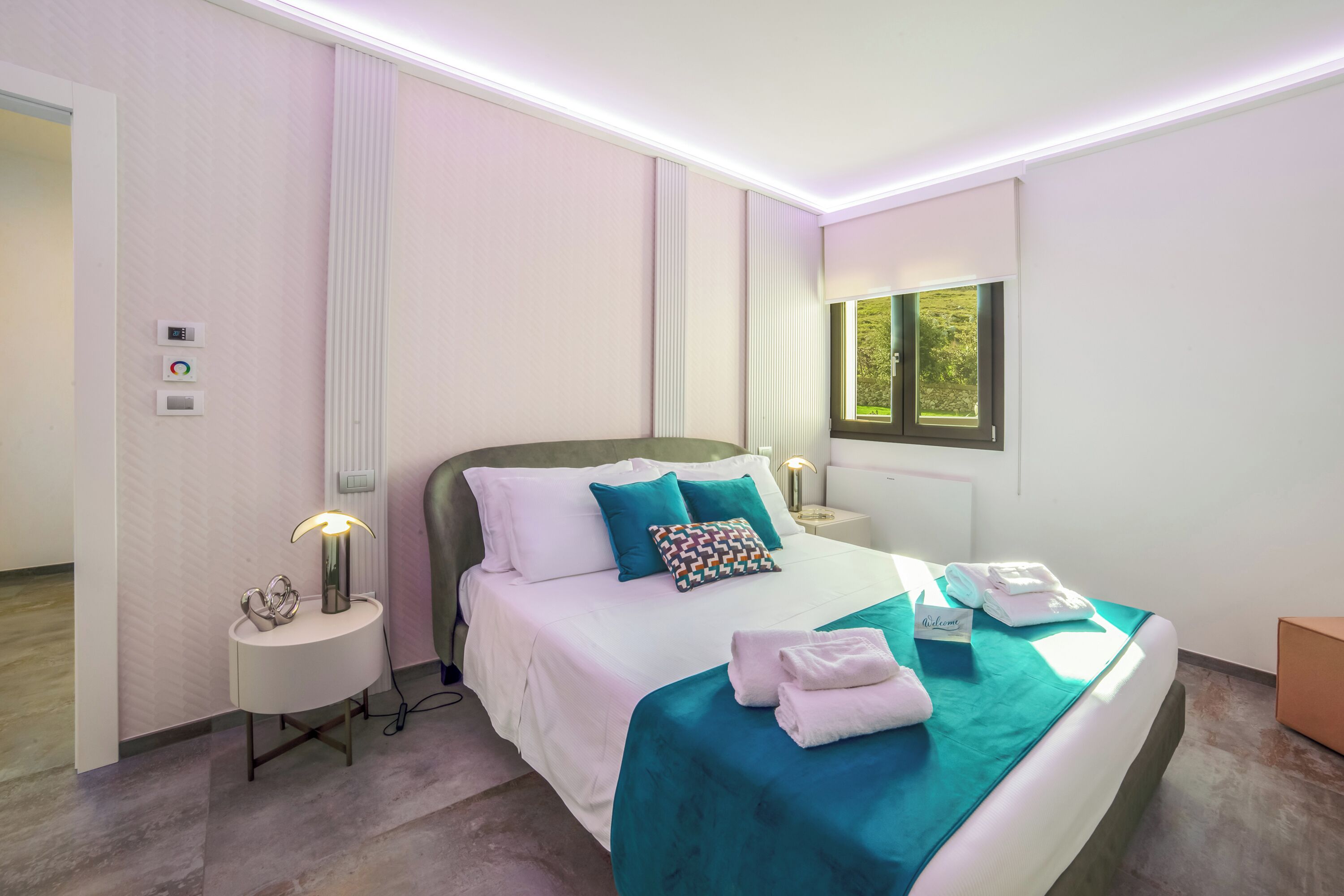 A modern hotel room with a neatly made bed, a bedside table with a lamp, a wall-mounted thermostat, and a window with a view of greenery.