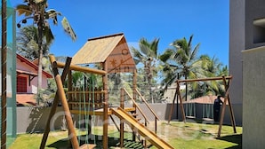 A playground with a slide and climbing structures, surrounded by palm trees and a building with a red roof. - Nomar Carneiros Ecoliving by CTA (Tamandaré)