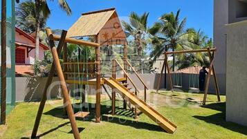 A playground with a slide and climbing structures, surrounded by palm trees and a building with a red roof.