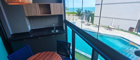 A balcony with a view of a swimming pool, a small kitchen area with a sink, and a wooden table and chair.
