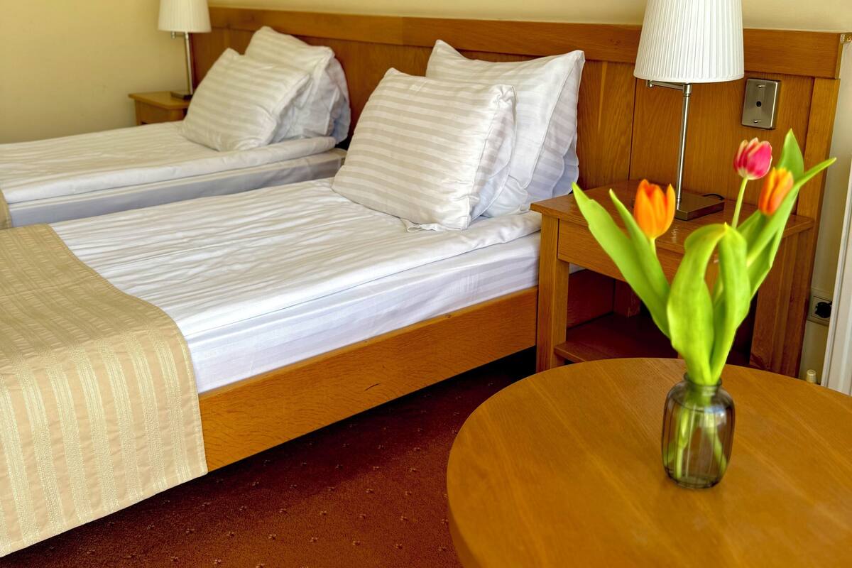 A hotel room with two beds, a wooden headboard, a bedside table with a lamp, a small table with a vase of flowers, and a painting on the wall.