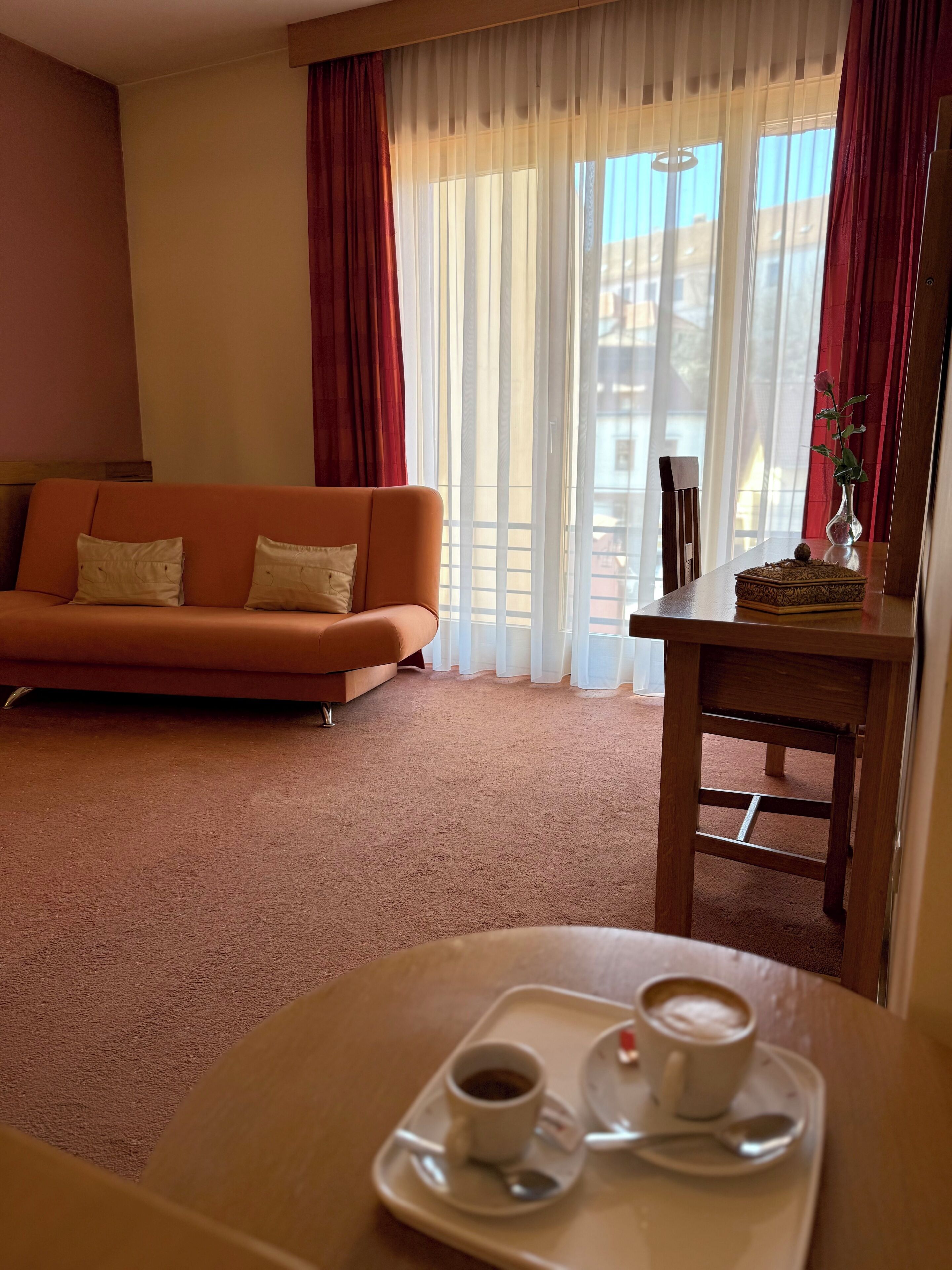 Una habitación con sofá, una mesa con una bandeja de café, un florero con una flor y una ventana con cortinas.