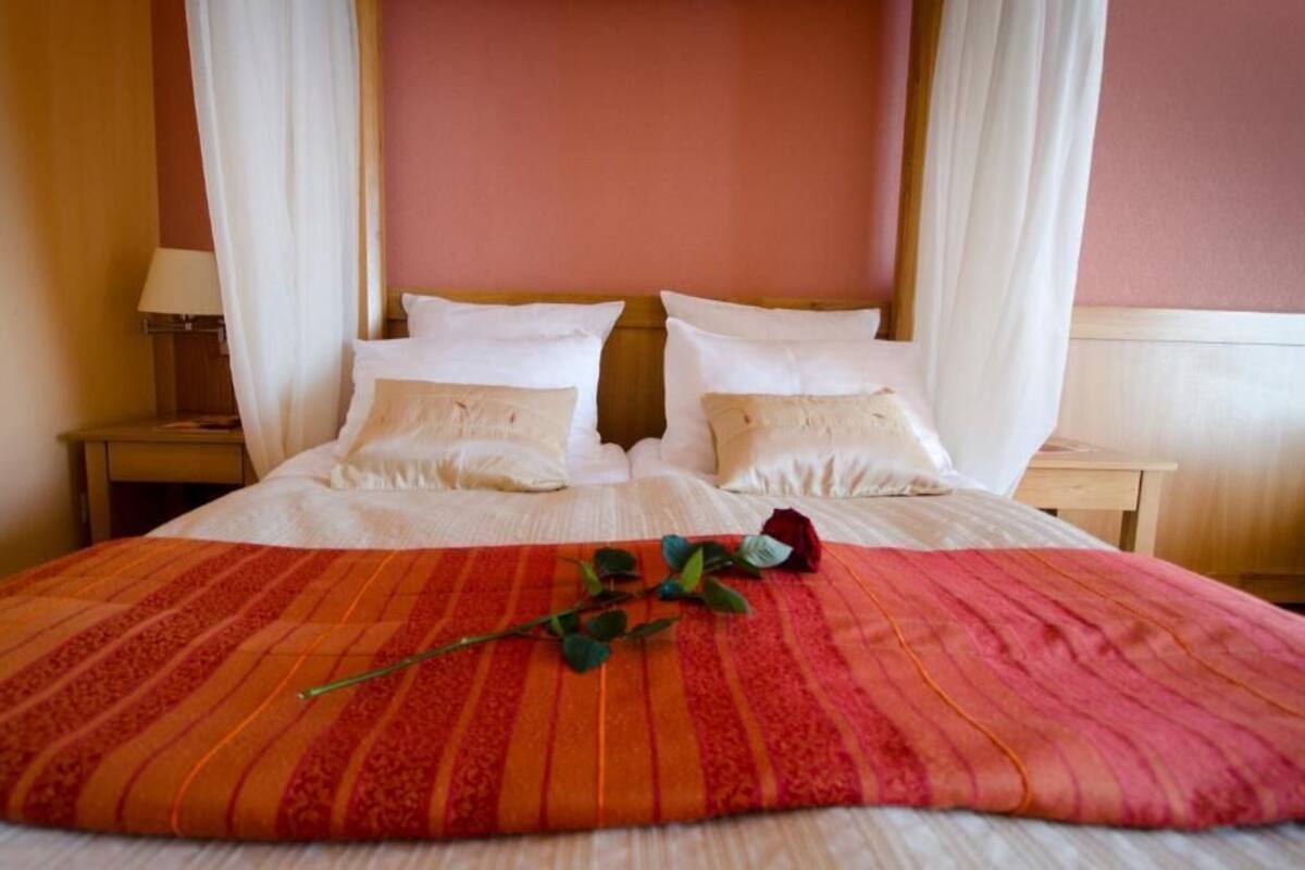A double bed with white linens, a red and orange striped blanket, and a single rose on the bed.