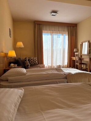 Une chambre d’hôtel avec deux lits, un bureau, une chaise, une lampe, une fenêtre avec des rideaux et un luminaire au plafond.