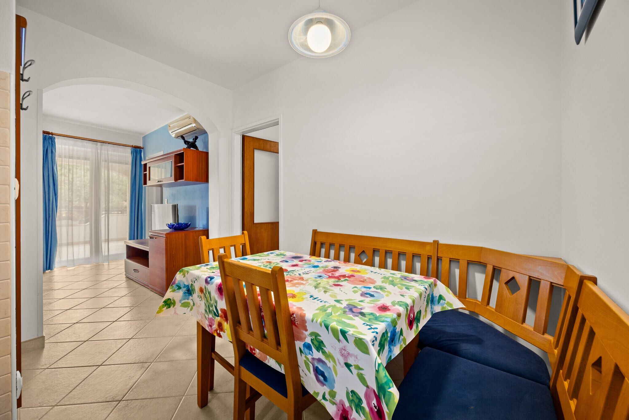 A dining area with a table covered in a floral tablecloth, wooden chairs, and a blue cushioned bench.