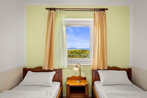 A room with two beds, a window with curtains, a nightstand with a lamp, and a view of greenery. - Family Apartment Stinjan with Sea View (Štinjan)