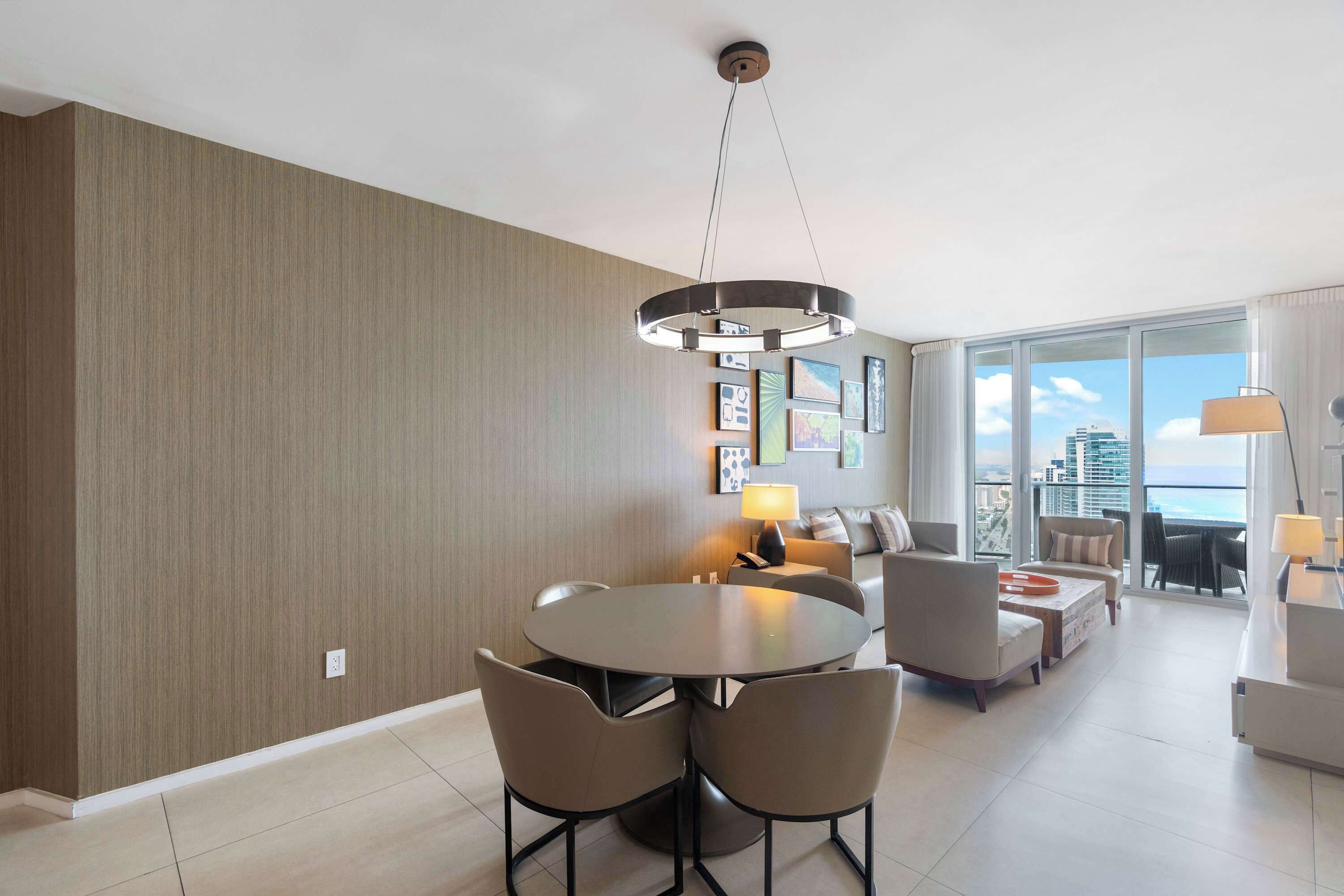 Una habitación de hotel moderna con zona de comedor, un conjunto de sofás, un amplio ventanal con vistas a la ciudad y una araña.