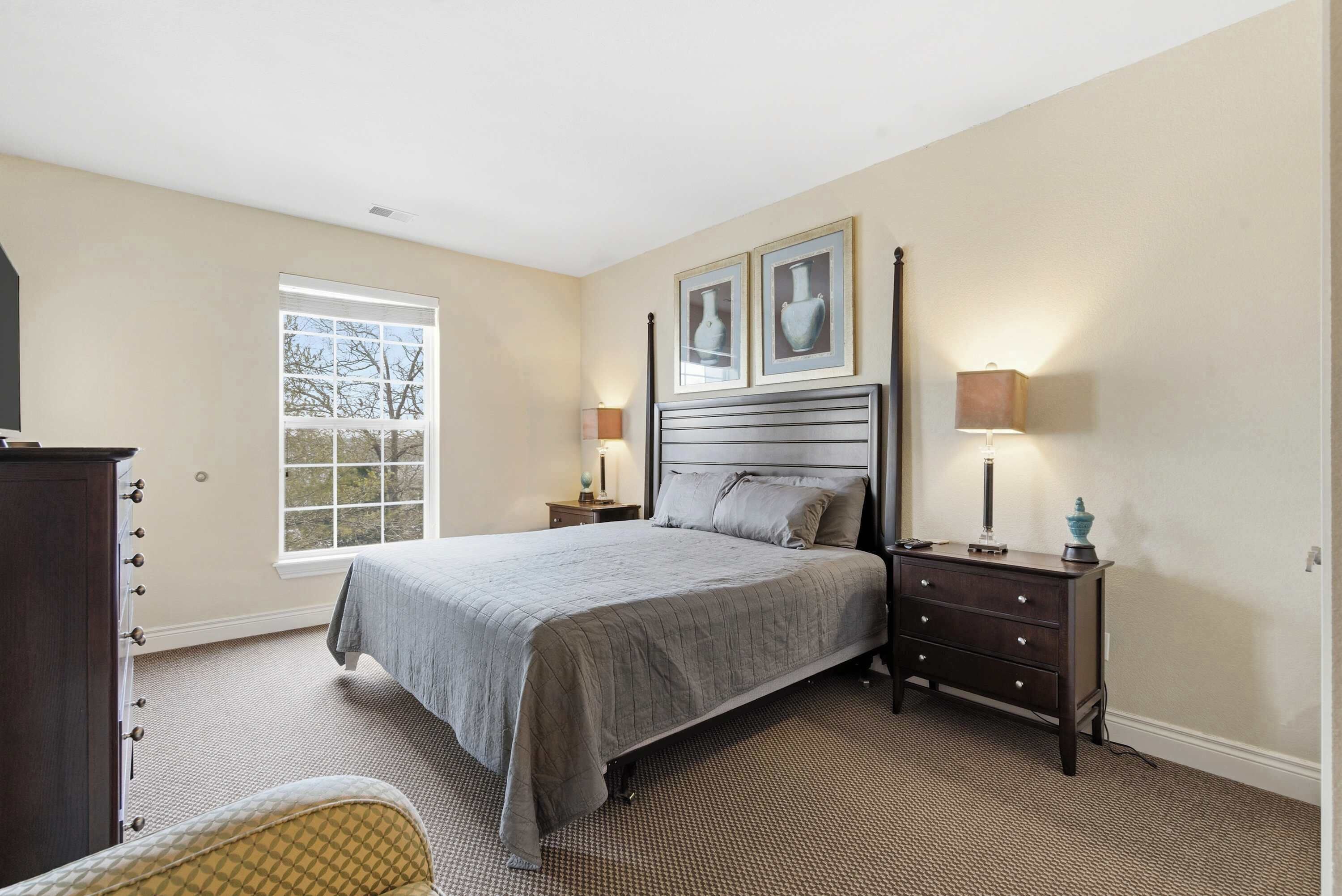 A bedroom with a large bed, a dresser, a nightstand, a lamp, and a window with a view of trees.