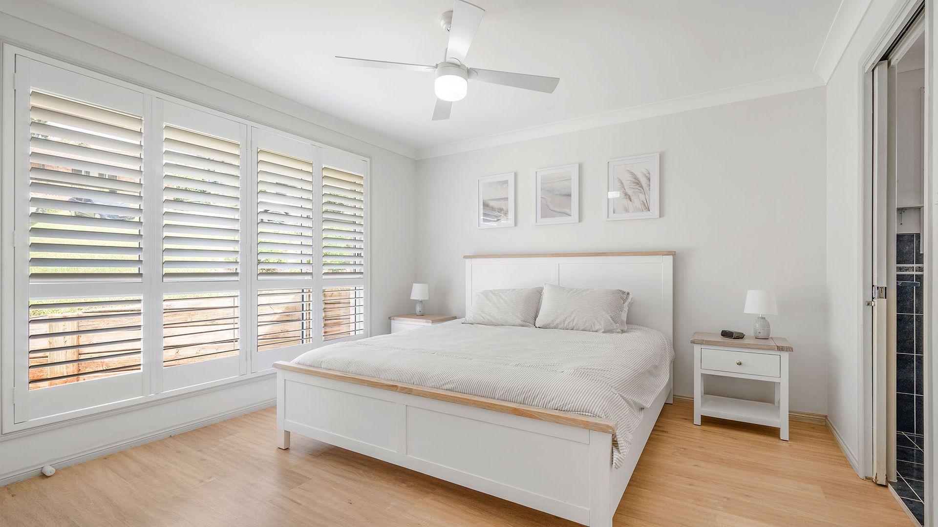 Un dormitorio con cama, mesitas de noche, ventilador de techo y ventanas amplias con persianas.