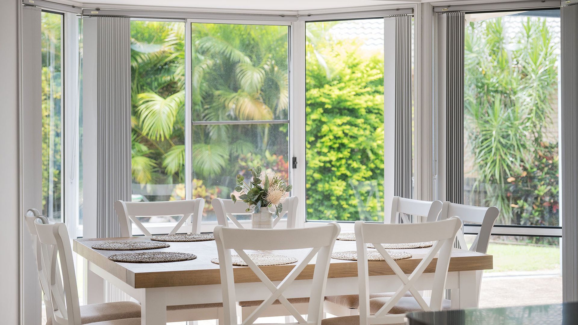 A dining area with a white table and chairs, a vase with flowers, and a view of greenery through large windows.