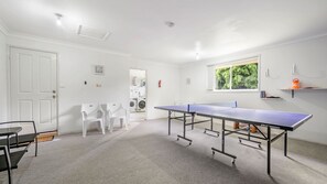 Ein Zimmer mit Tischtennisplatte, weißen Stühlen, einer Waschmaschine und einem Fenster mit Blick auf Bäume.