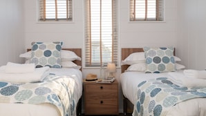 A twin bedroom with two beds, each with matching patterned bedding and pillows, a wooden nightstand between the beds, and a window with blinds. - Landal Bude Coastal Resort (Bude)