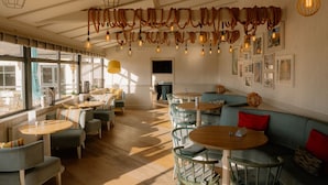 A spacious cafe with round tables and cushioned seating, large windows, and hanging light fixtures. - Landal Bude Coastal Resort (Bude)