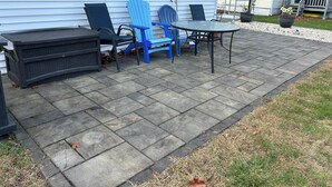 En udendørs terrasse med et bord, stole, en lille sort bænk og en potteplante.
