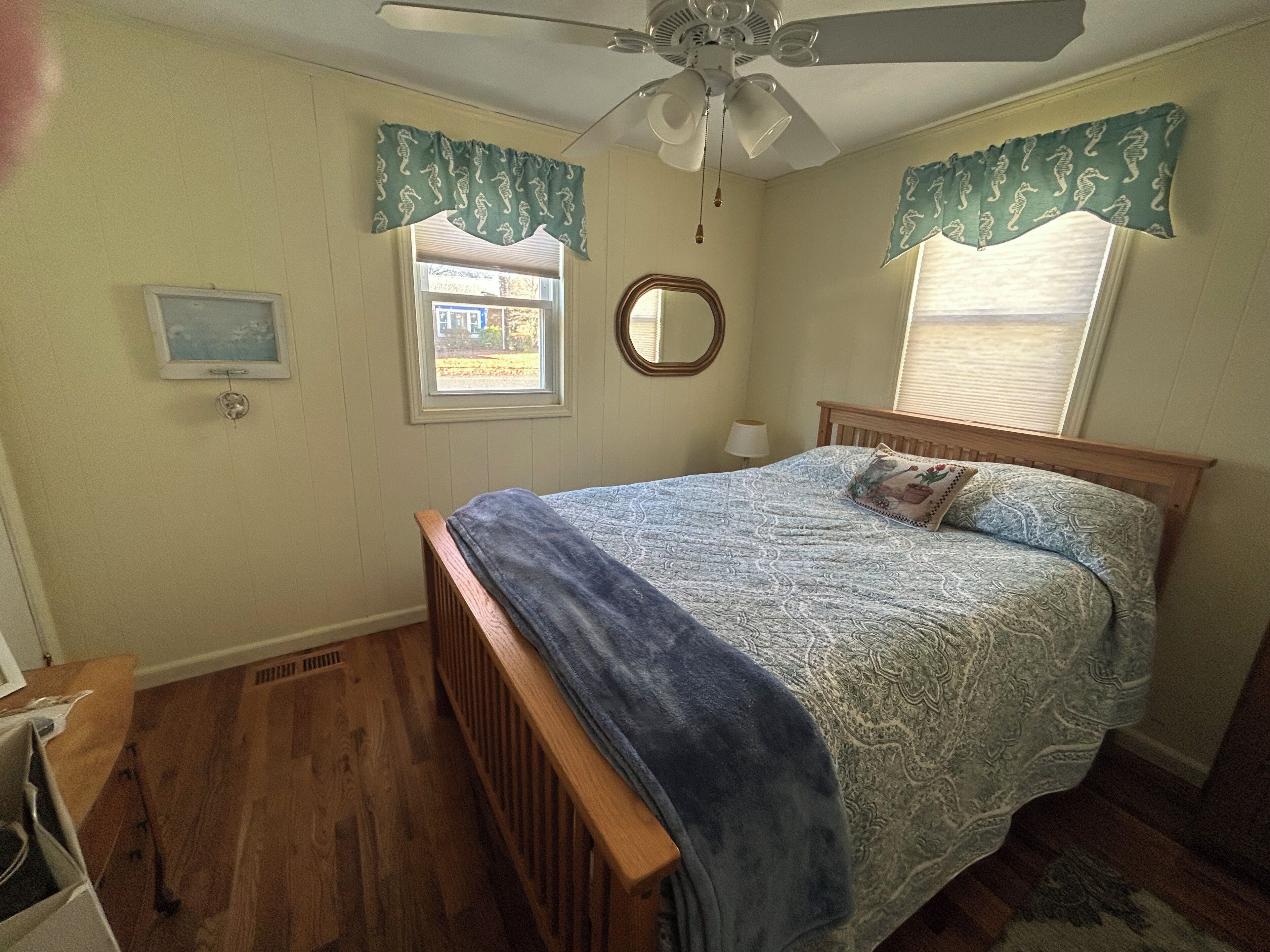 A bedroom with a wooden bed, a ceiling fan, a window with shutters, a mirror, and a small window with a view of the outside.