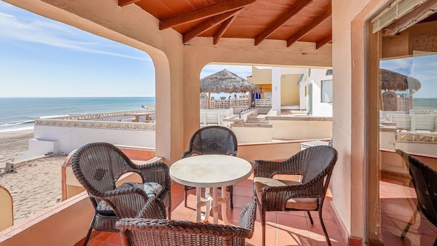 A covered outdoor seating area with wicker chairs and a table, overlooking a beach and a thatched-roof structure. - Casa Puesta del Sol – Beachfront, Rocky Point (Puerto Peñasco)